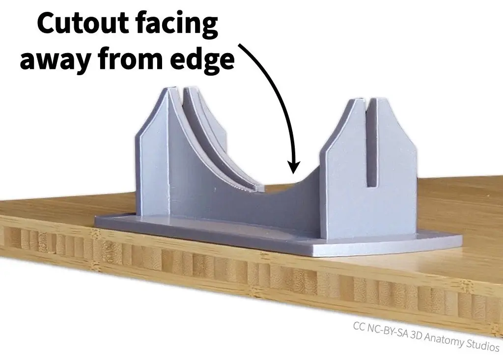 Photo of the cross-section plate bracket sitting at the edge of a tabletop with text and an arrow indicating that the cutout faces away from the edge